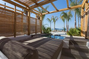 A sunlit outdoor lounge area with a pool, wooden pergola, and palm trees. - Branco Mykonos (Mykonos)
