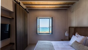 A bedroom with a large bed, a TV, a window with a view of the sea, and wooden elements. - Branco Mykonos (Mykonos)