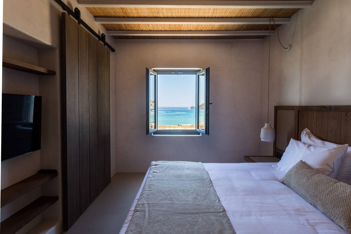 A bedroom with a large bed, a TV, a window with a view of the sea, and wooden elements.