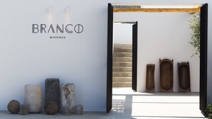 A minimalist entrance with a wooden beam ceiling, a white wall with the text "BRANCO MYKONOS", and a series of stone and wooden objects on the ground.