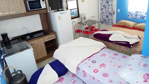Una habitación compacta con kitchenette, una cama con ropa de cama floral, una mesita y una silla.