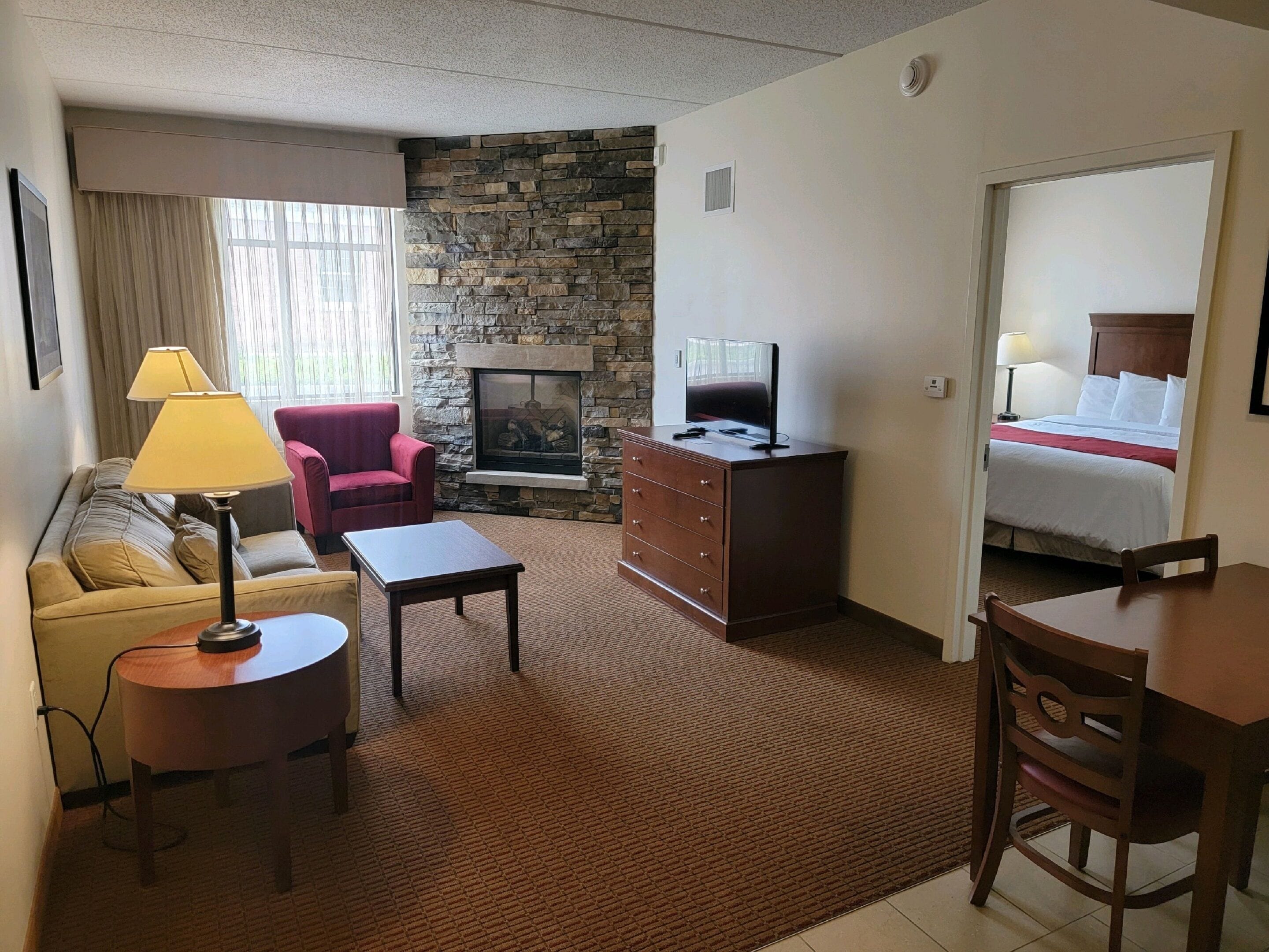 Une chambre d’hôtel avec une cheminée en pierre, un canapé, un fauteuil rouge, une commode en bois, un téléviseur posé sur le dessus, et un lit avec une literie blanche.