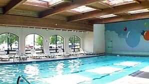 Una piscina interior con agua azul cristalina, techo iluminado por el sol y sillas de descanso blancas.
