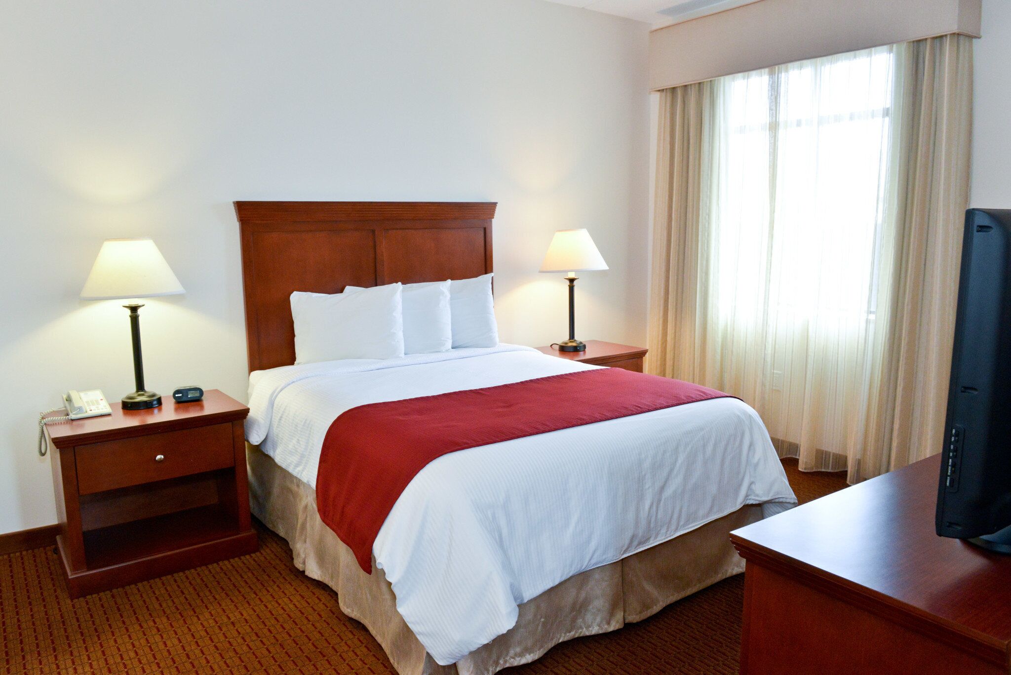 A hotel room with a large bed, two bedside tables with lamps, a TV on a wooden desk, and a window with curtains.