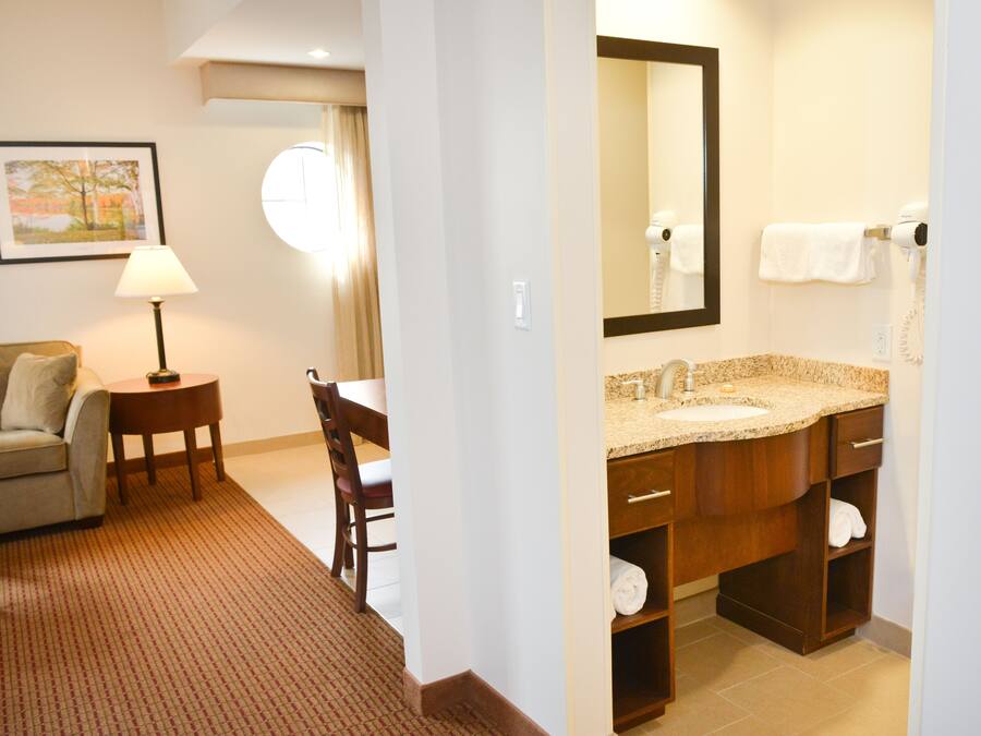 Une chambre d’hôtel comprenant un lit, un bureau, une chaise, une lampe, une salle de bain avec un lavabo et un miroir, et un tableau encadré au mur.