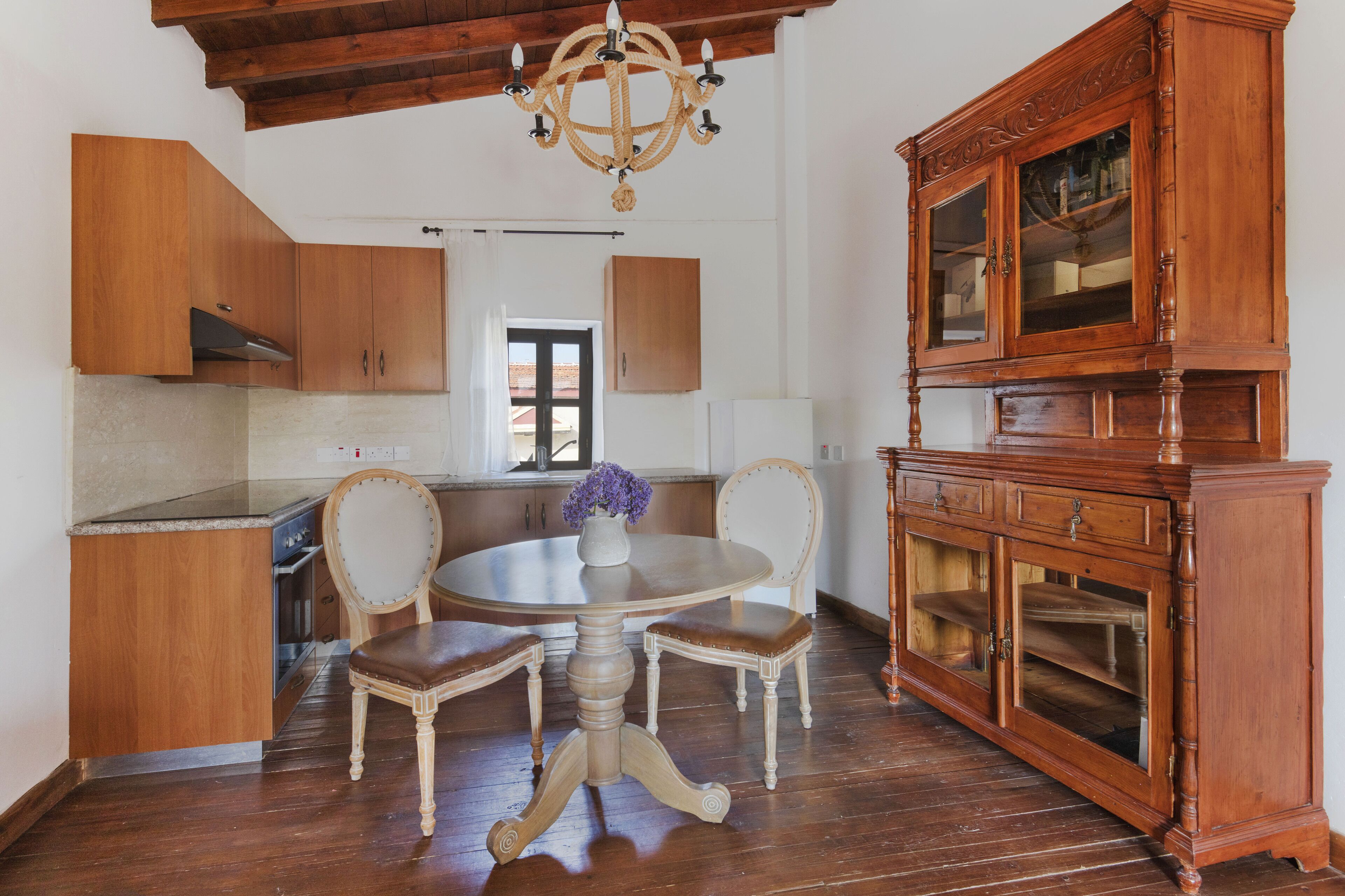 Una cocina con armarios de madera, una mesa de comedor con sillas, una lámpara de araña y un aparador de madera con puertas de vidrio.