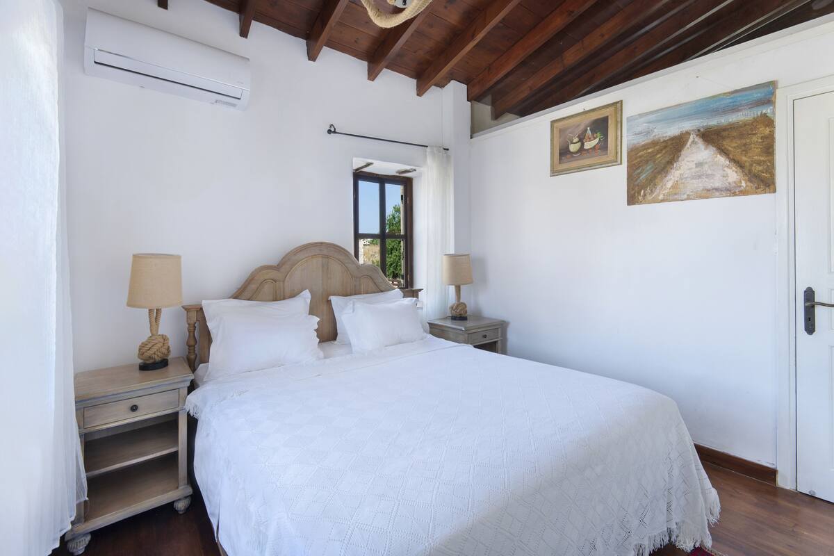 A bedroom with a large bed, two bedside tables with lamps, a wooden headboard, a window with a view, and two paintings on the wall.