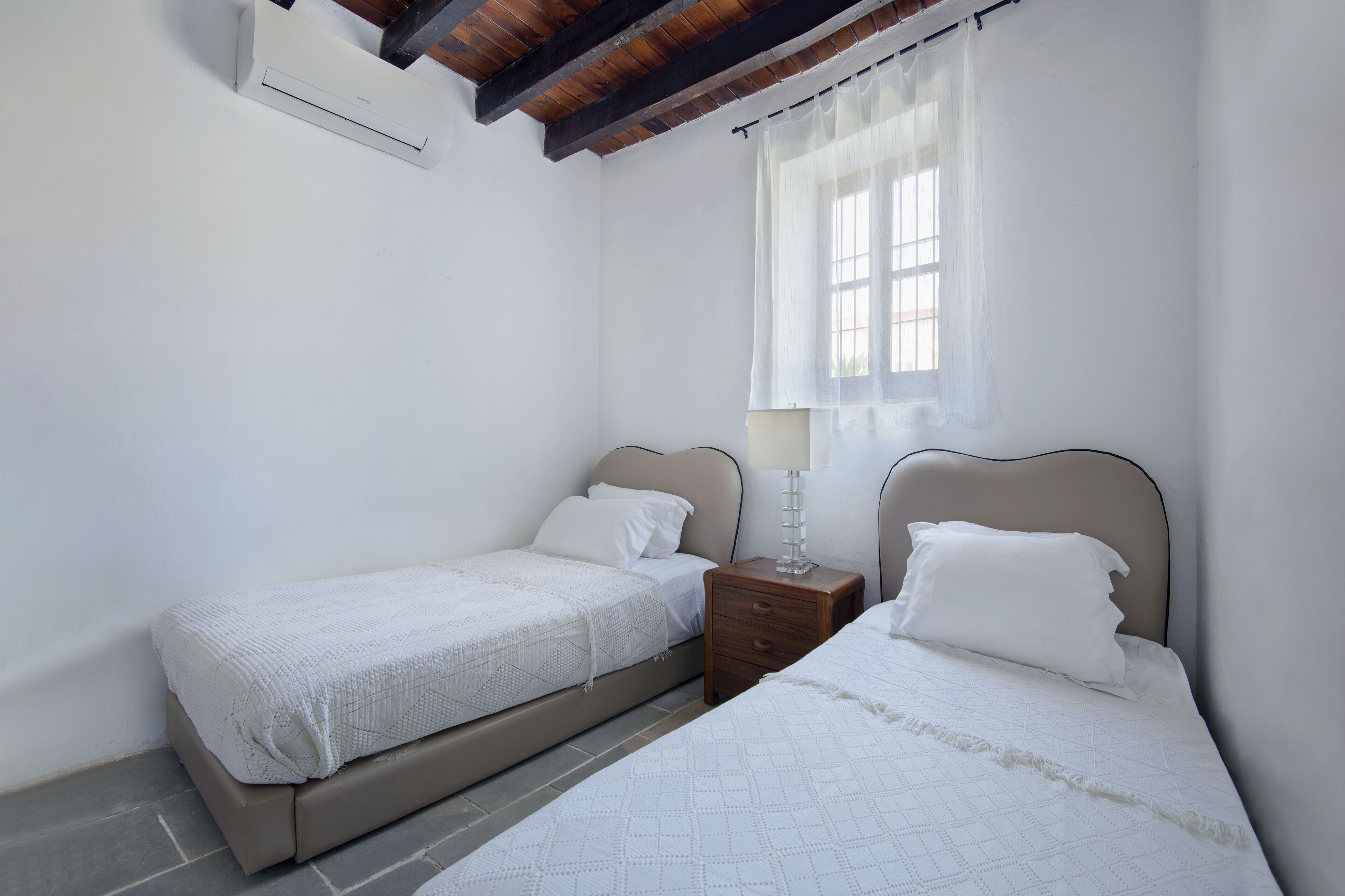 A bedroom with two beds, a window with sheer curtains, a bedside table with a lamp, and a ceiling fan.