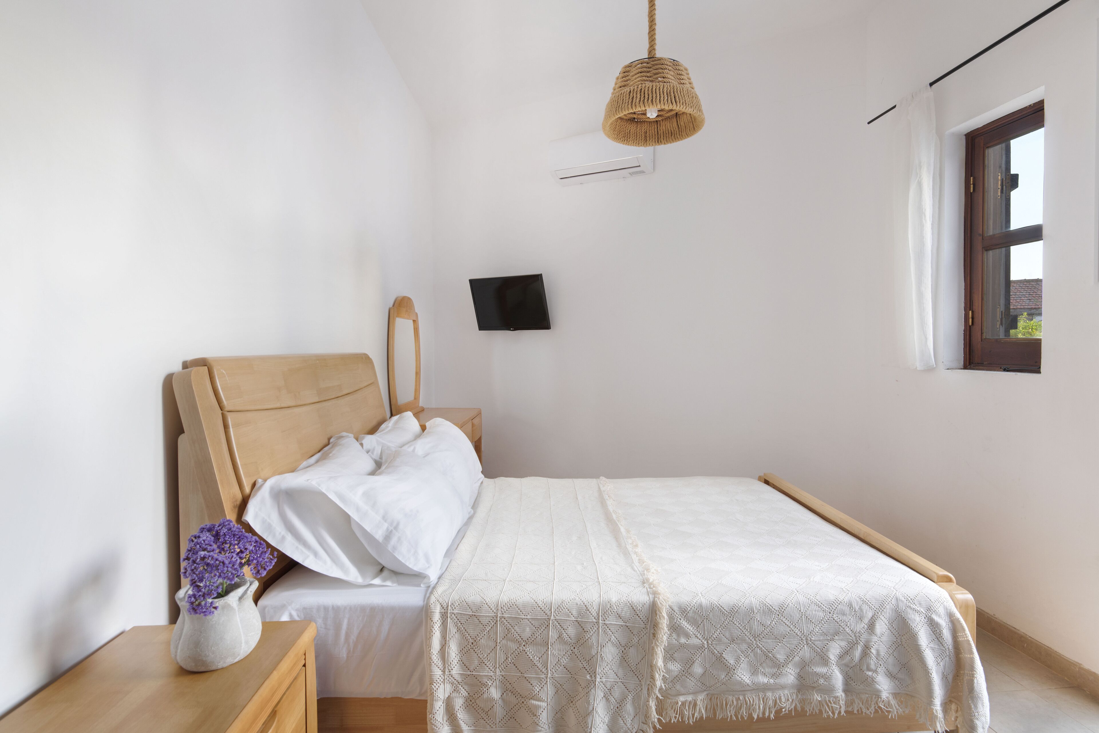 Une chambre à coucher avec un lit en bois, une table de chevet ornée d’un vase de fleurs, une lampe suspendue et une fenêtre.