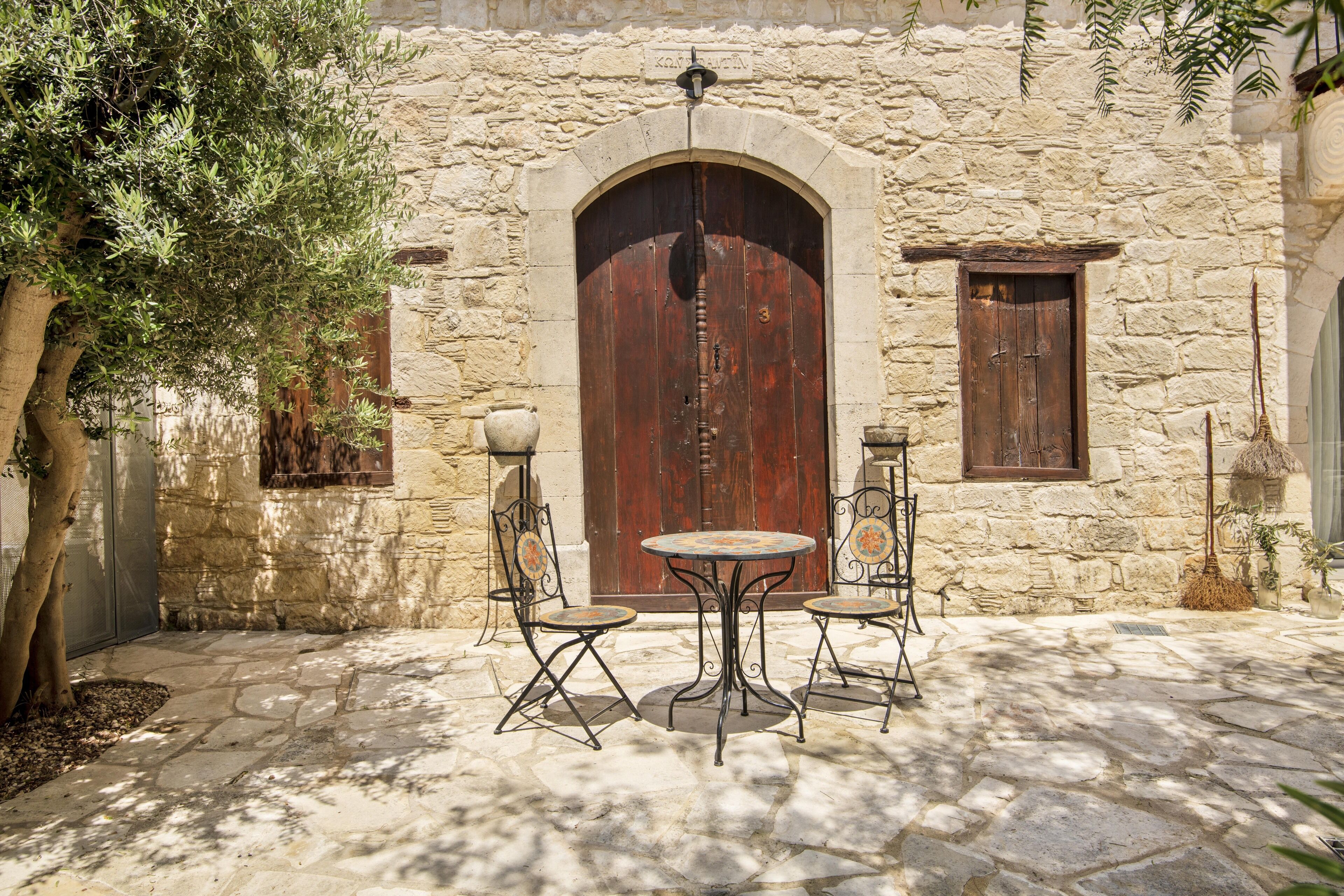 A stone building with a large wooden door, two metal tables with chairs, and a tree.
