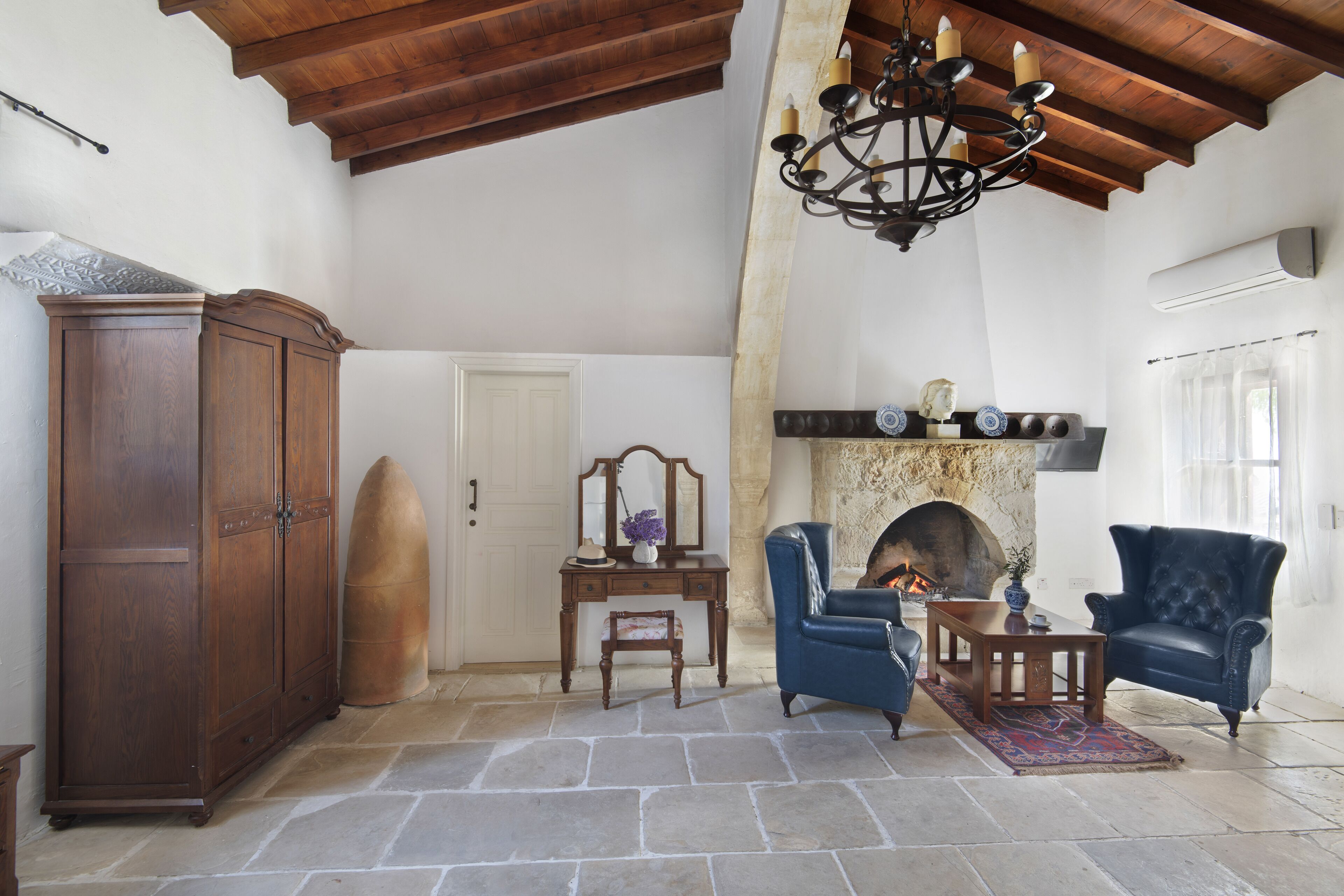 A spacious living room with a stone floor, wooden furniture, a fireplace, and a high wooden ceiling.