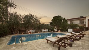 A swimming pool with lounge chairs and tables, surrounded by trees and a building. - Meleni Cottage Houses (Pareklisia)