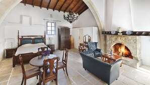 A spacious room with a large bed, a fireplace, a dining area, and a living area with a blue sofa. - Meleni Cottage Houses (Pareklisia)