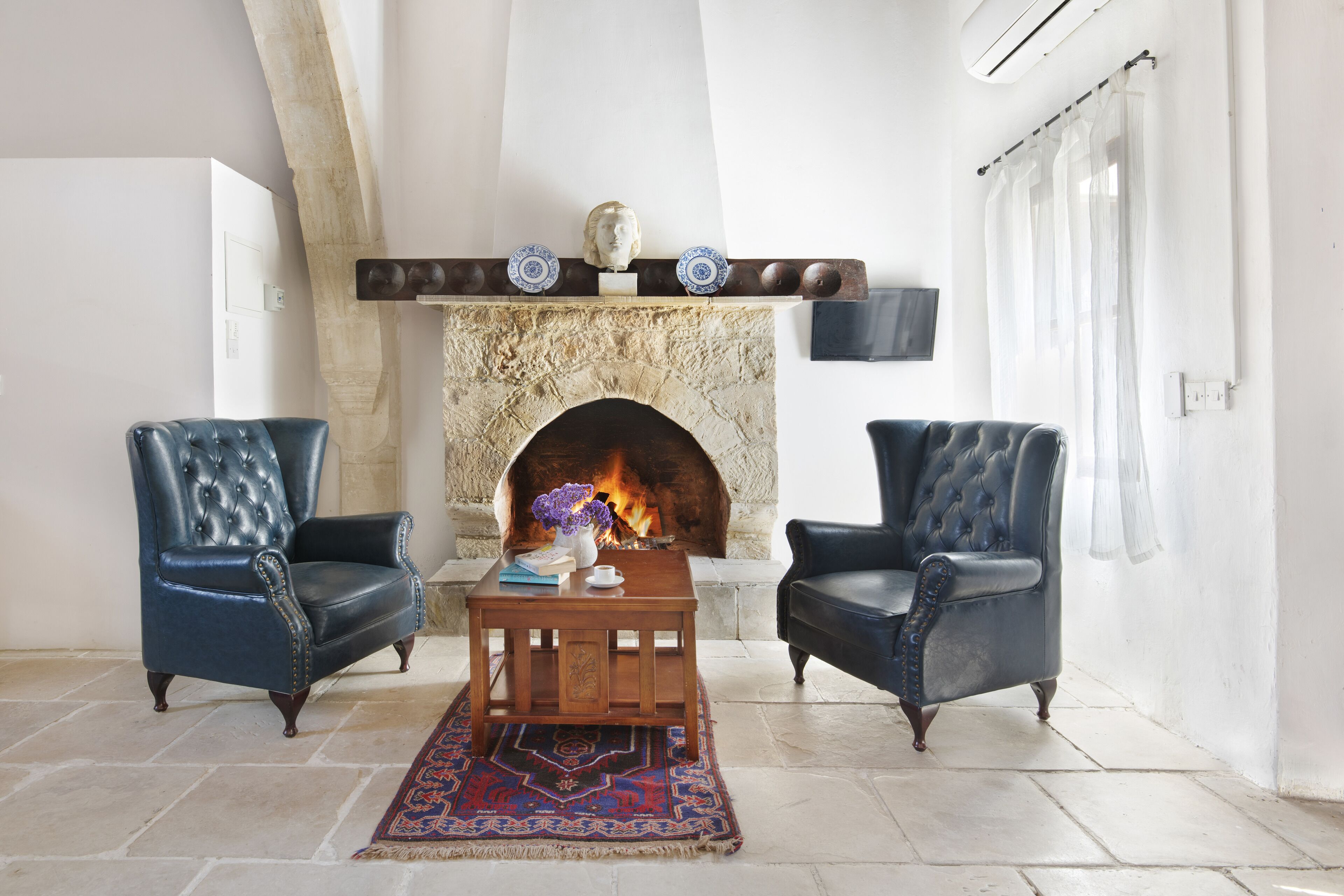 A living room with two leather armchairs, a wooden coffee table, a patterned rug, a fireplace with decorative plates, and a mounted television.