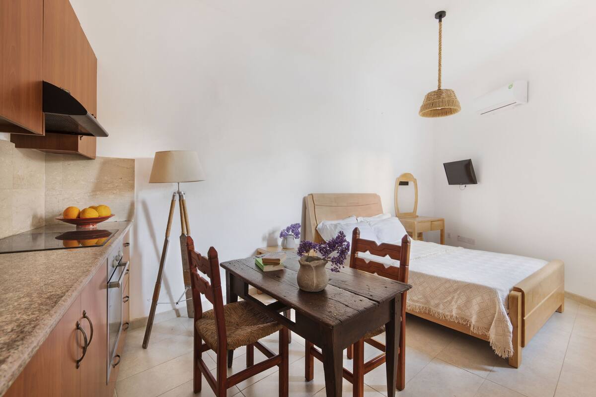 A compact bedroom with a bed, bedside table, lamp, and a small table with a vase and books.