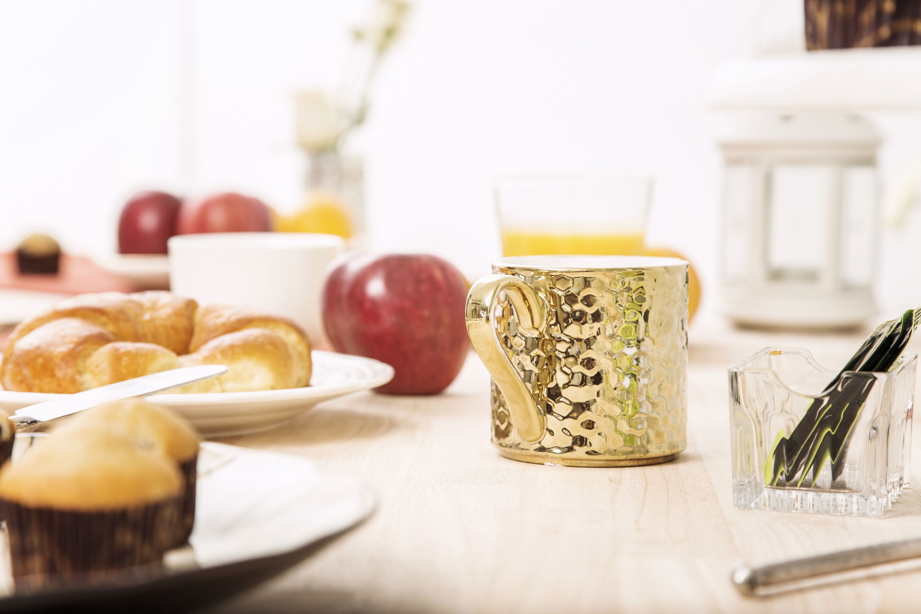 A breakfast table with croissants, apples, a coffee pot, and a glass of orange juice.