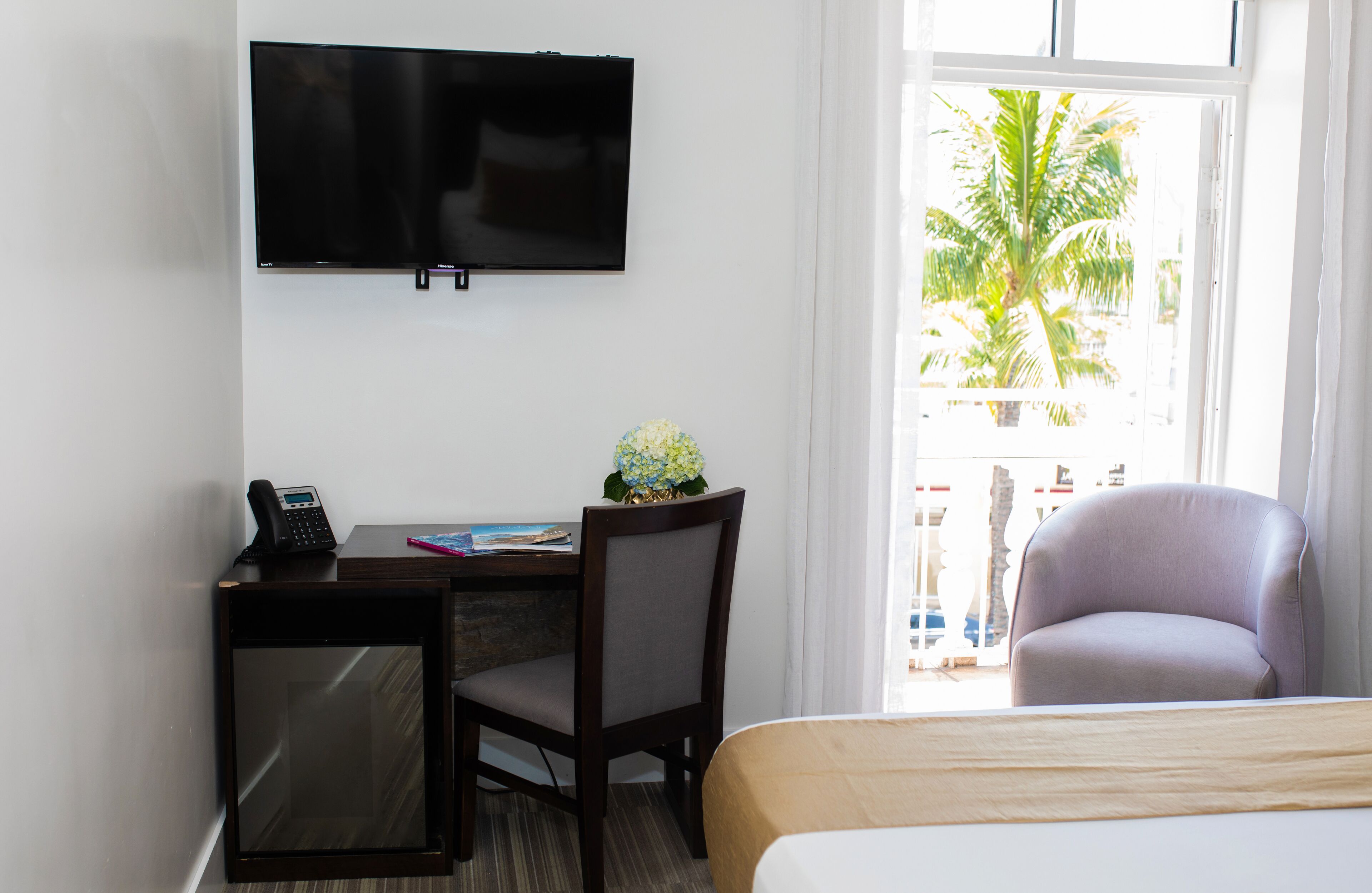 Habitación de hotel con cama, escritorio con computadora, una silla, televisor montado en la pared y ventana con vista a palmeras.