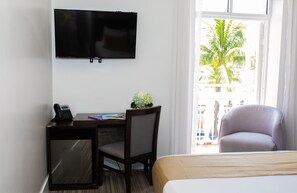 A hotel room with a bed, a desk with a computer, a chair, a TV mounted on the wall, and a window with a view of palm trees. - Casa Boutique Hotel (Miami Beach)