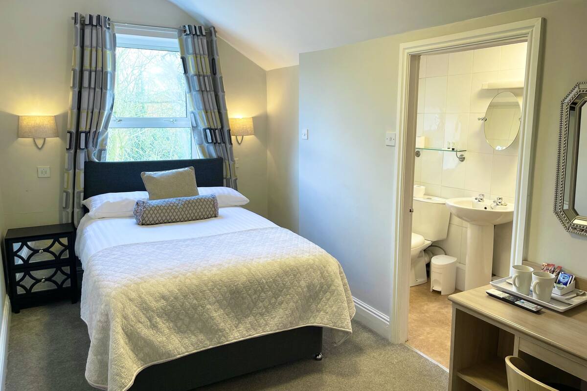 A hotel room with a bed, a bedside table, a window with curtains, a bathroom with a sink and mirror, and a tray with cups and a remote control.