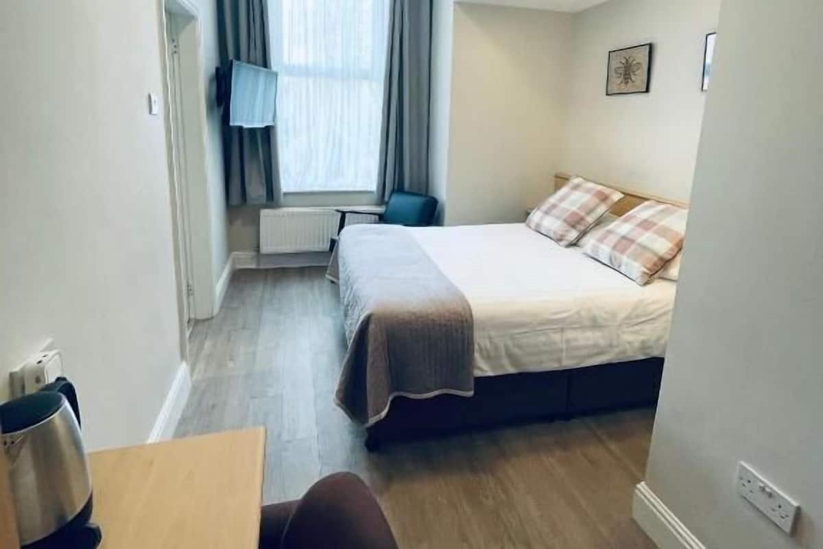 A compact hotel room with a bed, a desk, a chair, a radiator, and a window with curtains.