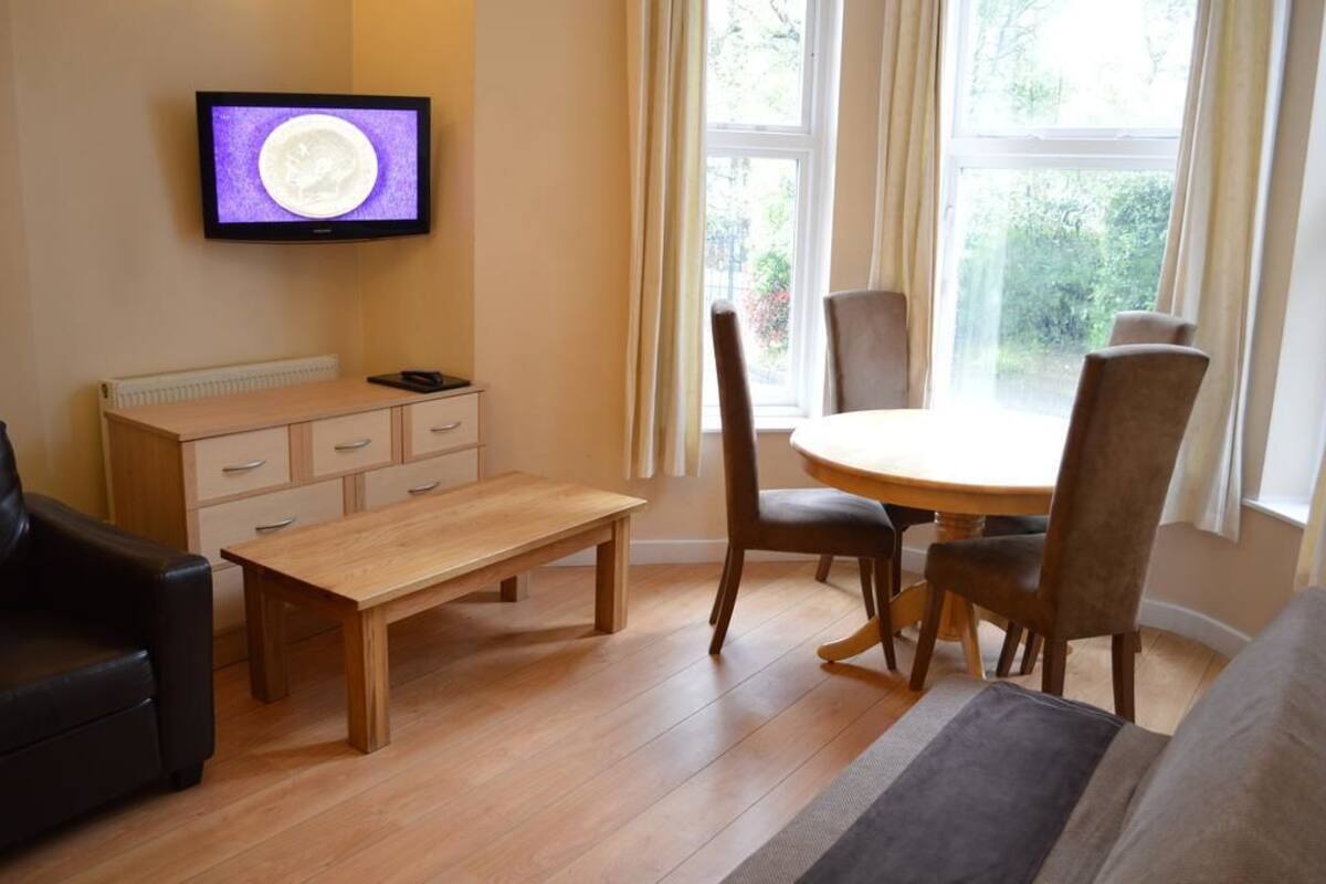 A living room with a wooden table, two chairs, a sofa, a TV mounted on the wall, and a window with curtains.