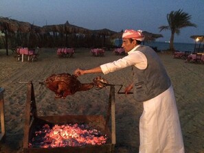 Una persona asando carne en un asador sobre fuego directo en la playa.