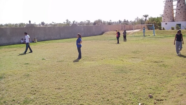A grassy field with a few people walking, a soccer goal, and two tall structures in the background. - Six Corners Resort (Fayed)
