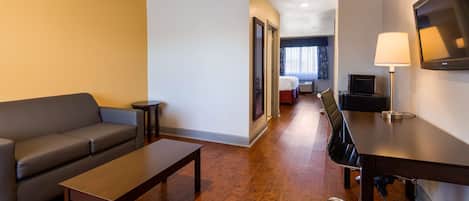 A hotel room with a sofa, a small table, a desk with a chair, a TV, and a lamp.