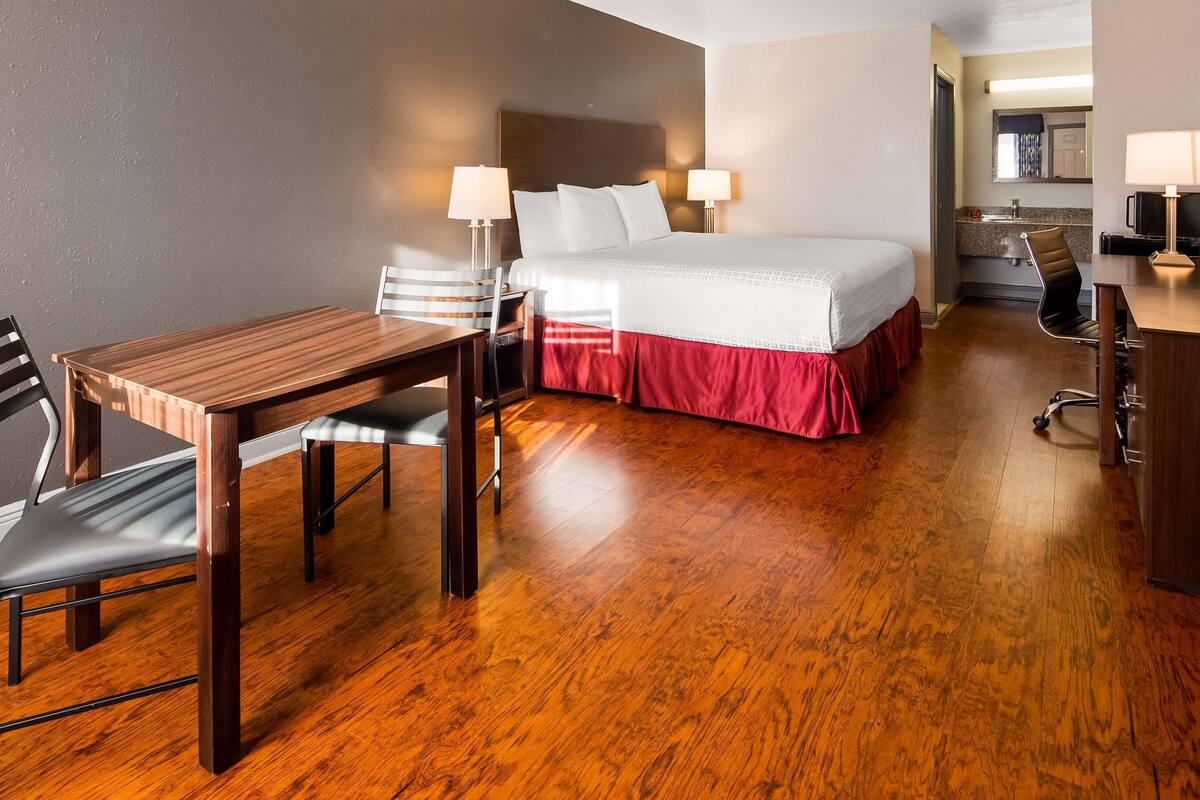 Une chambre d’hôtel avec un plancher en bois, un lit avec une jupe rouge, un bureau avec une chaise et un téléviseur fixé au mur.