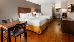 Une chambre d’hôtel avec une table à manger en bois, deux lits avec du linge de lit blanc, un bureau avec une lampe et un téléviseur fixé au mur.