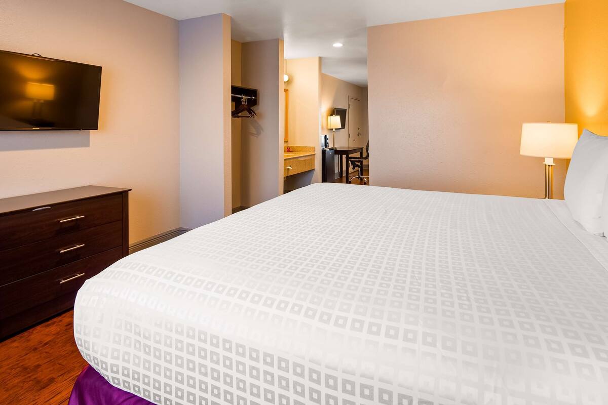 Une chambre d’hôtel avec un grand lit, une commode, un téléviseur fixé au mur et une salle de bain visible à travers une porte ouverte.