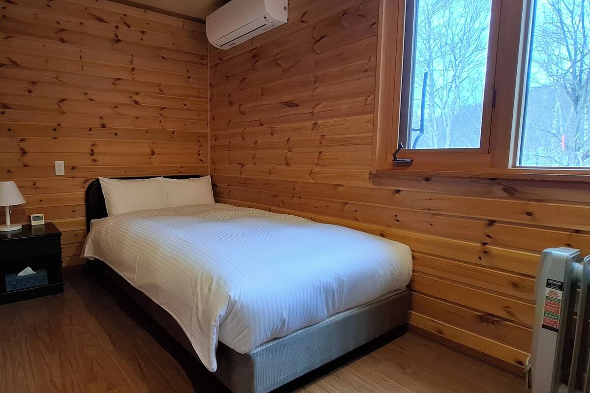 A small, cozy room with wooden walls, a single bed, a nightstand with a lamp, and a window with a view of trees.