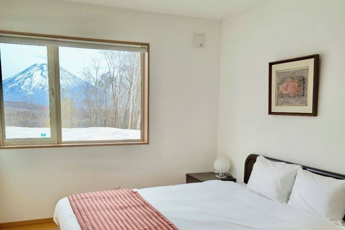 A bedroom with a bed, a window with a mountain view, a framed picture on the wall, and a nightstand with a lamp.