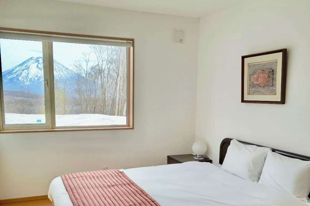 Un dormitorio con cama, ventana con vista a la montaña, un cuadro en la pared y mesita de noche con lámpara.