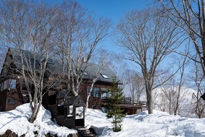 Una cabaña de madera con techo inclinado, rodeada de nieve y árboles sin hojas.