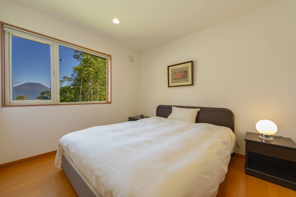 A bedroom with a large bed, a nightstand, a bedside table with a lamp, a window with a view of a mountain, and a framed picture on the wall.