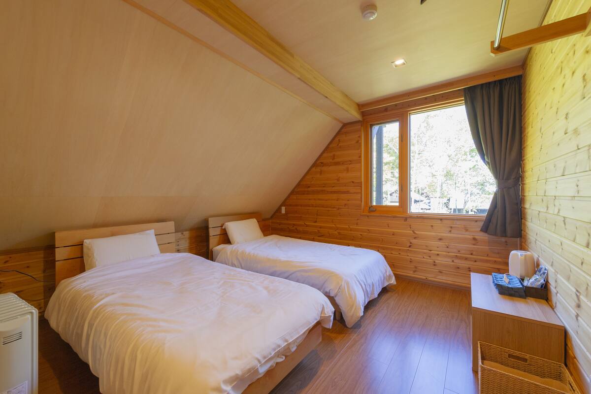 A bedroom with two beds, wooden walls, a window with curtains, and a small table with a lamp.