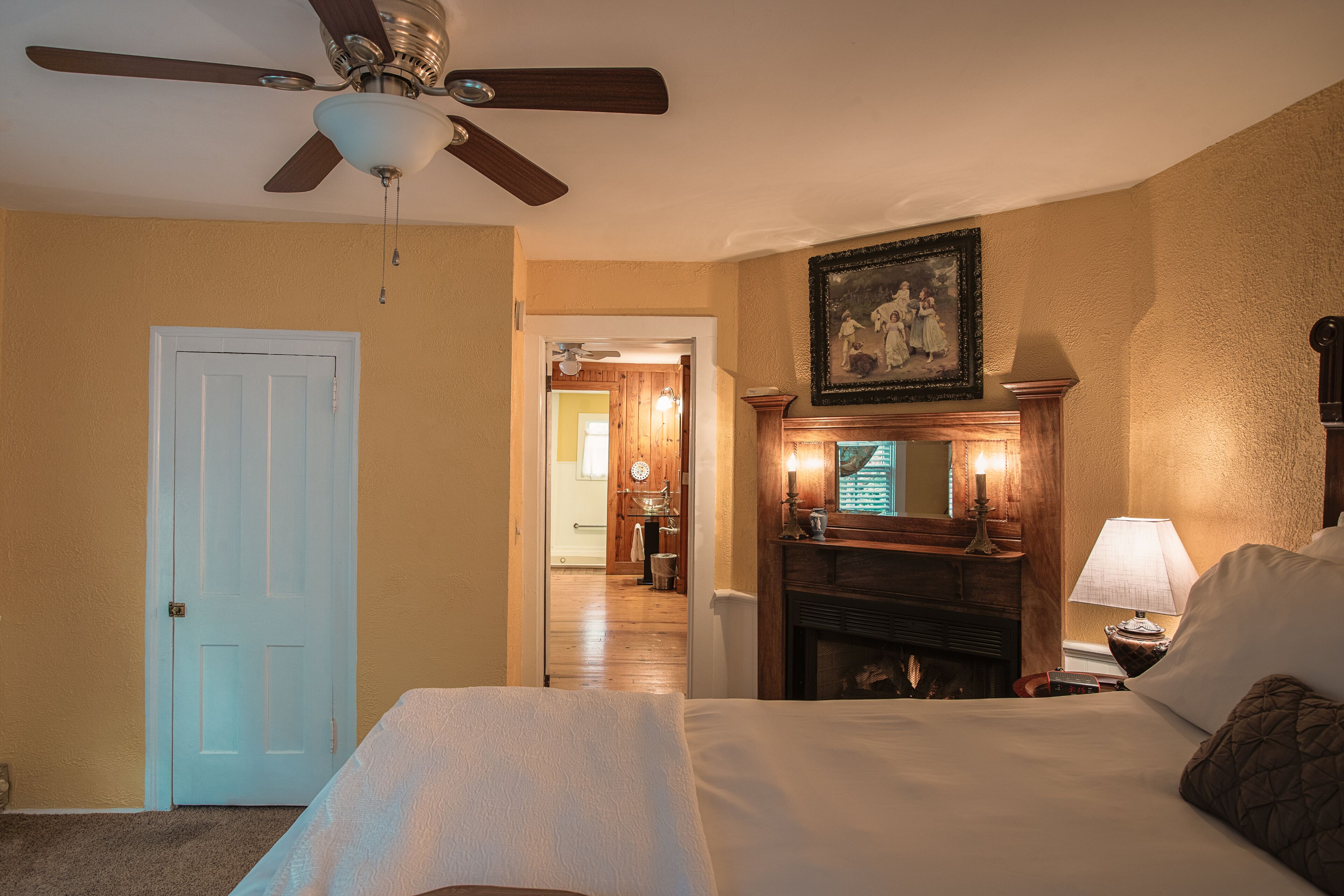 Habitación de hotel con cama, ventilador de techo, chimenea y un cuadro en la pared.
