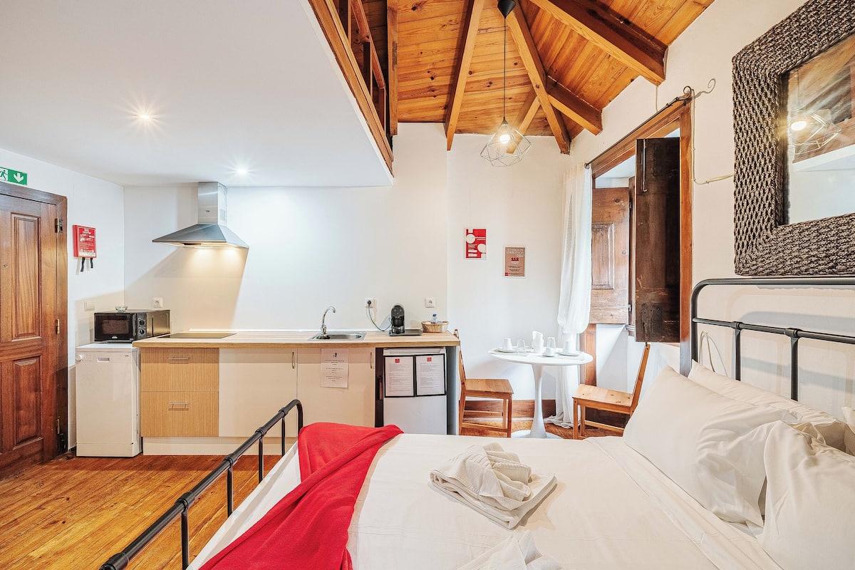 A compact room with a bed, a small kitchenette, a dining table, and a wooden ceiling.