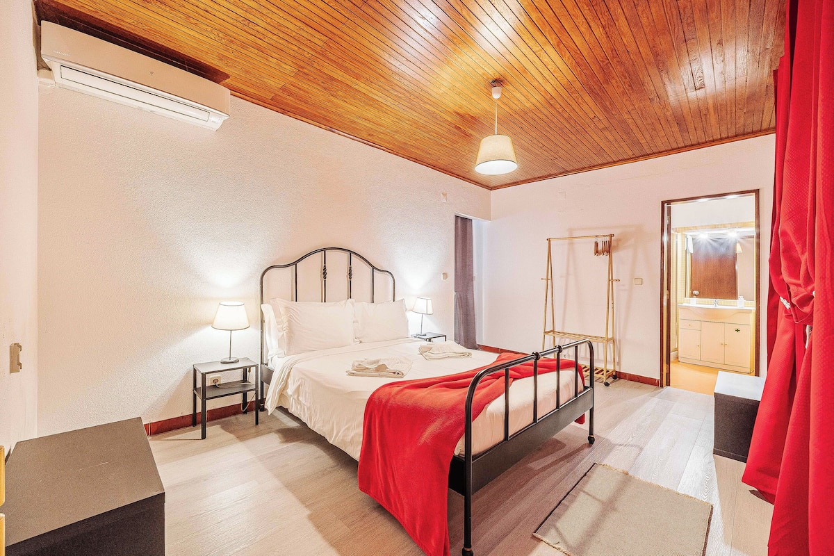 A bedroom with a wooden ceiling, a bed with a red blanket, bedside lamps, and a visible bathroom door.