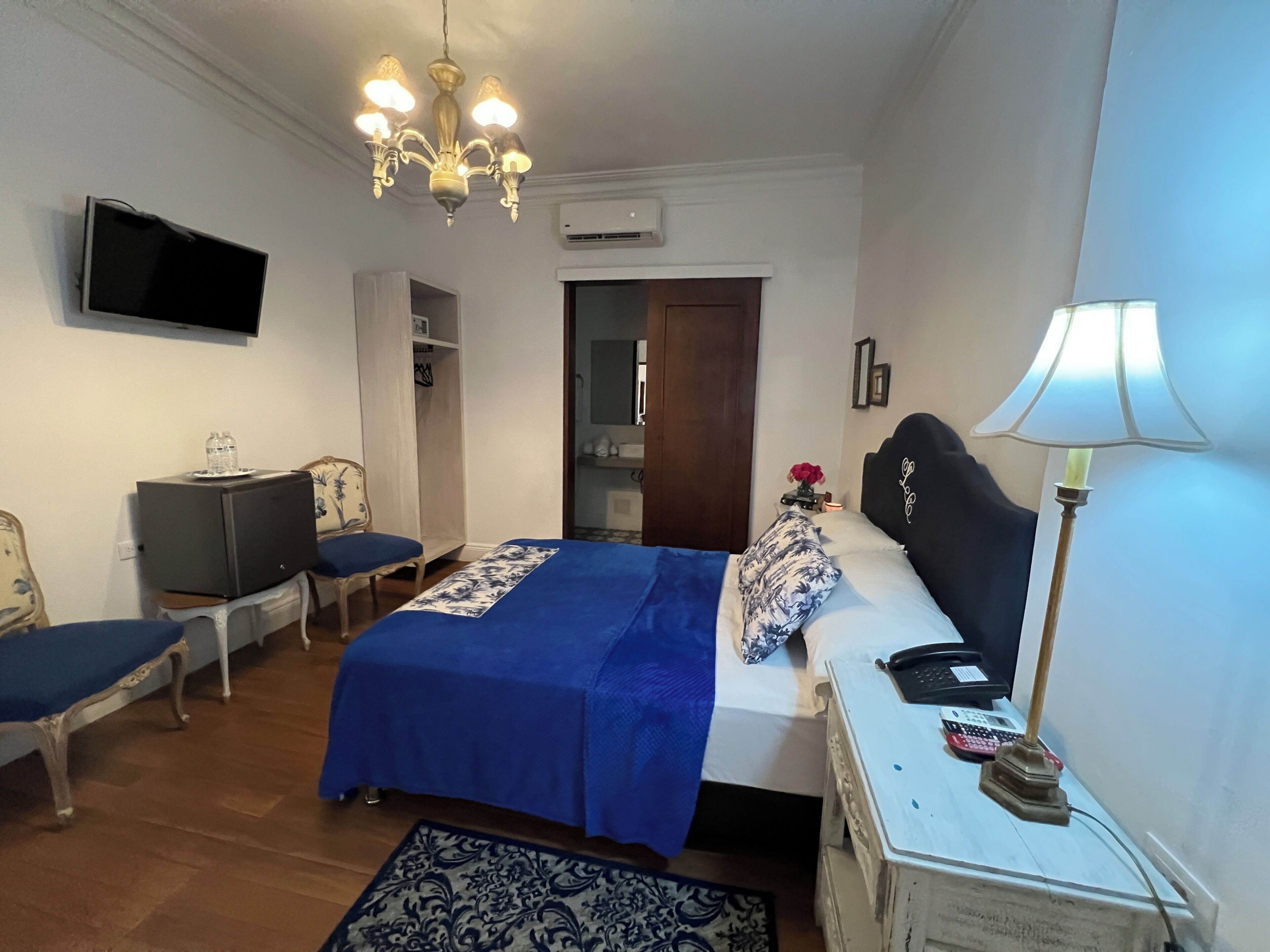 A bedroom with a bed, a desk, a chair, a TV, a chandelier, and a lamp.