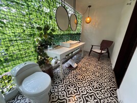 Un baño con inodoro, lavabo y silla, con baldosas de suelo estampadas y una pared cubierta de follaje verde.