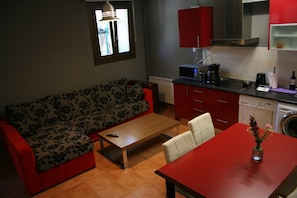 A compact living space with a sofa, a small dining table, a coffee machine, and a washing machine. - Alojamientos Acá y Allá - Hostel (Esteribar)