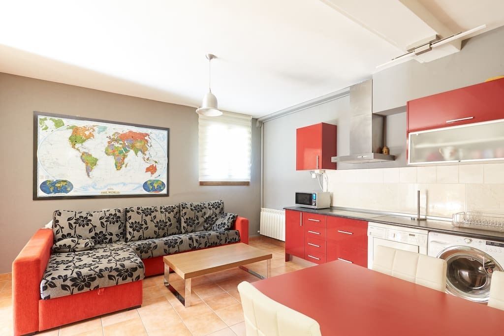 A compact living space with a sofa, a small dining table, a kitchen area with red cabinets, a washing machine, and a world map on the wall.