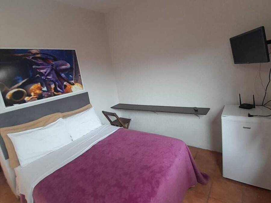 Un dormitorio con cama, televisión de pantalla plana, un mueble blanco pequeño y un cuadro en la pared.