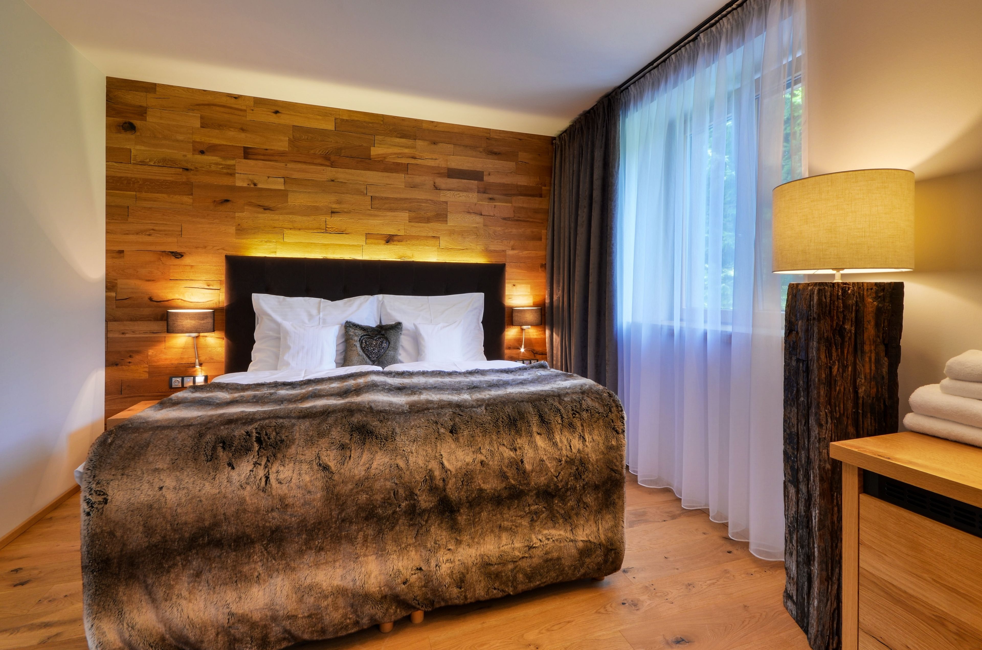 Un dormitorio moderno con cabecera de madera, una colcha mullida y grande, dos lámparas de noche y vistas a un bosque a través de cortinas traslúcidas.