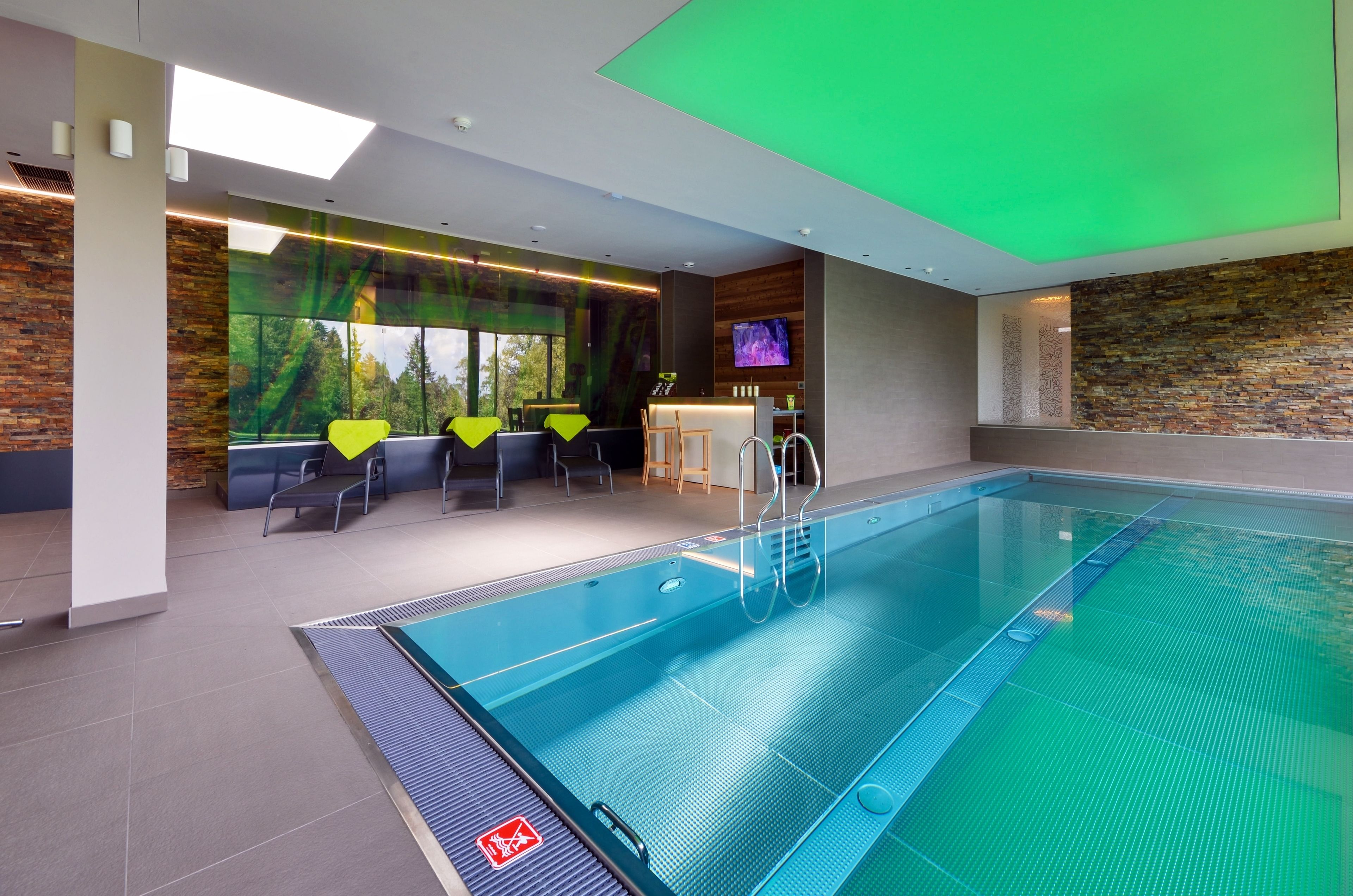 Una piscina interior moderna con vistas despejadas al exterior a través de amplios ventanales, una zona de recepción con bar y un área de descanso con sillas amarillas.