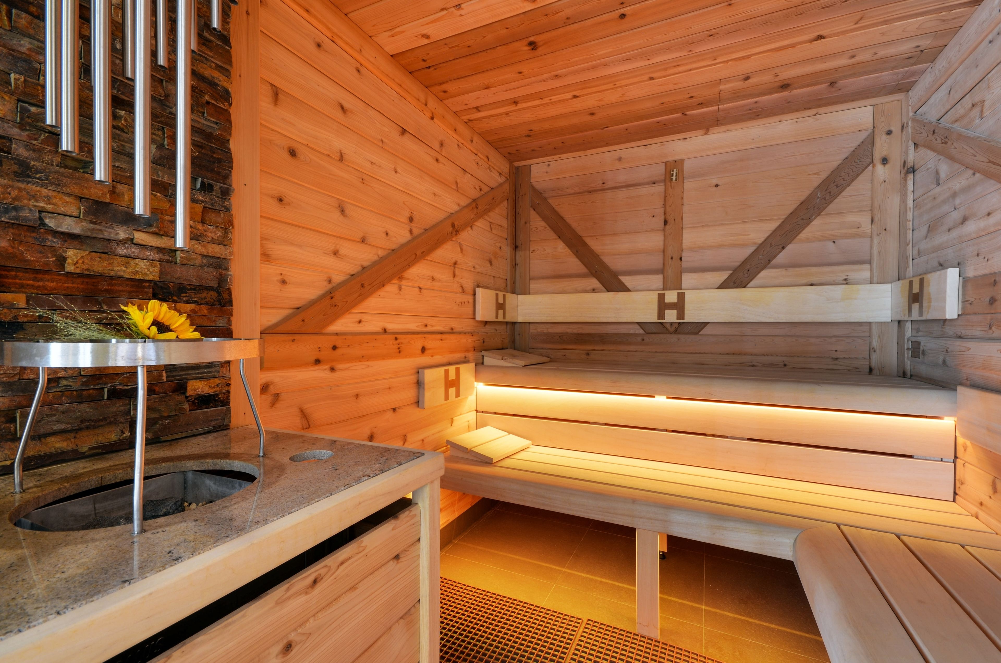 A sauna with wooden walls and benches, a stone wall with a metal grate, and a small table with a flower.