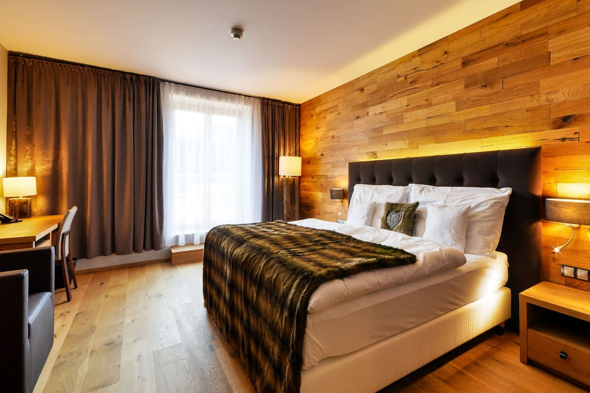 Habitación de hotel con cabecera de madera, cama grande con ropa blanca, escritorio con silla y ventana con cortinas.