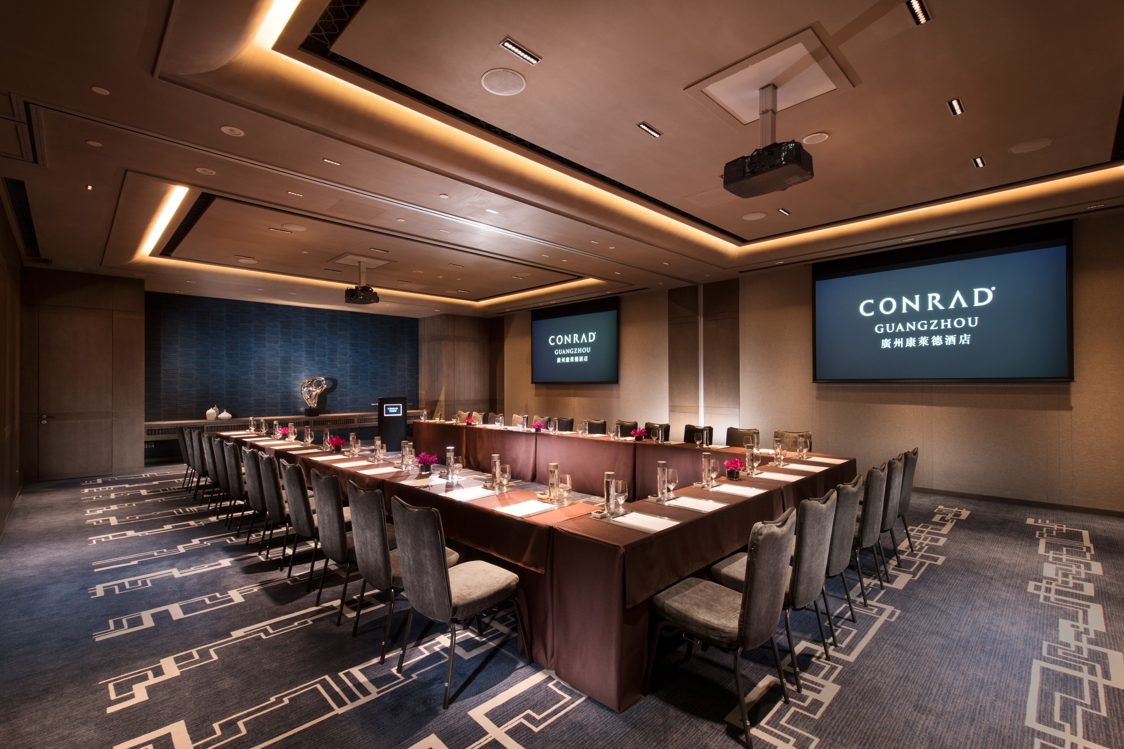 Un salón de conferencias con una larga mesa, sillas y una pantalla proyectora mostrando 'CONRAD GUANGZHOU'.